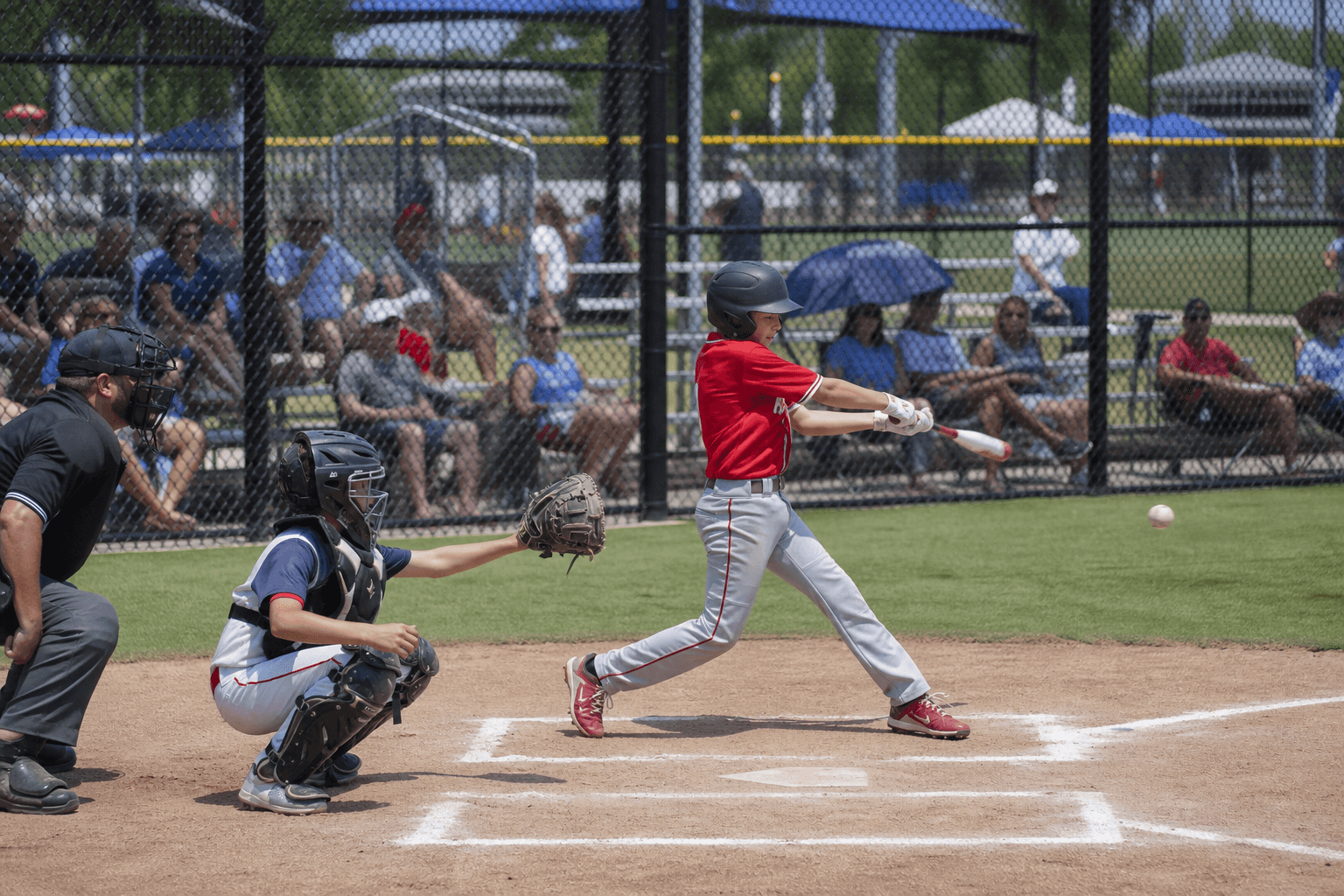 Youth travel baseball batter mid-swing at home plate during a weekend tournament with parents watching from bleachers along the fence