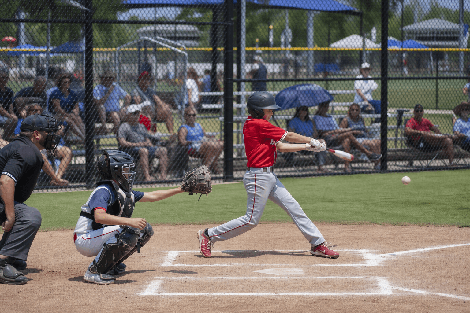 Youth travel baseball batter mid-swing at home plate during a weekend tournament with parents watching from bleachers along the fence