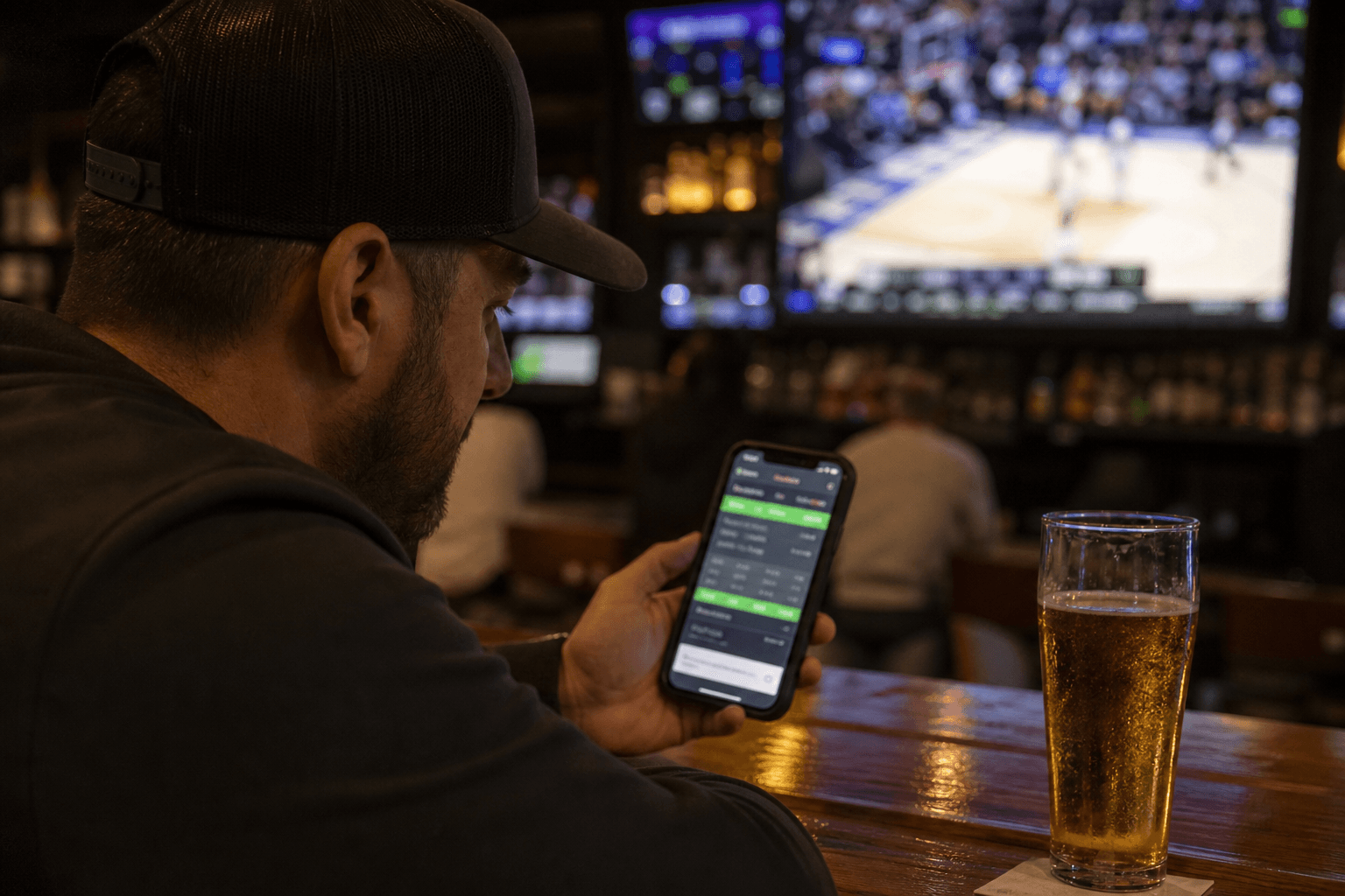 Man at a sports bar counter checking a sports betting app on his smartphone while a basketball game plays on the TV behind the bar